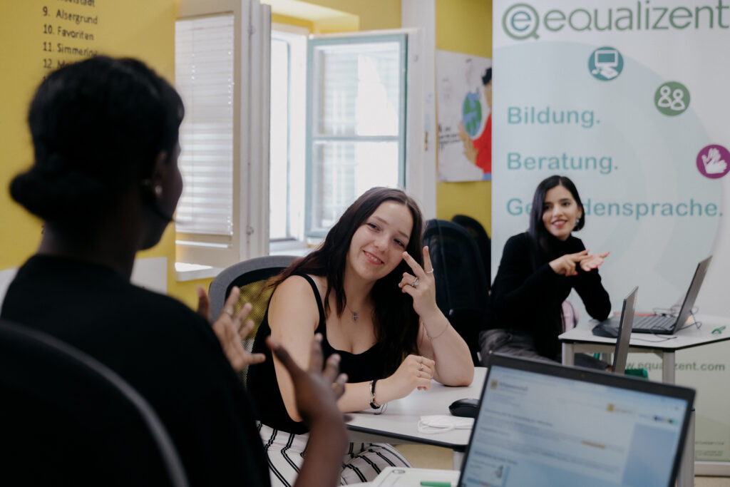 3 gehörlose Teilnehmer_innen in einer equalizent Schulung gebärden. Hintergrund: equalizent Rollup mit Text "Bildung. Beratung. Gebärdensprache."
